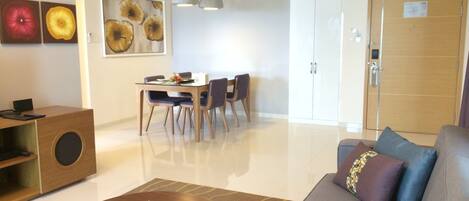 A modern living room with a sofa, coffee table, dining table, and chairs. There is a cabinet with a speaker, artwork on the walls, and a door with a number sign.