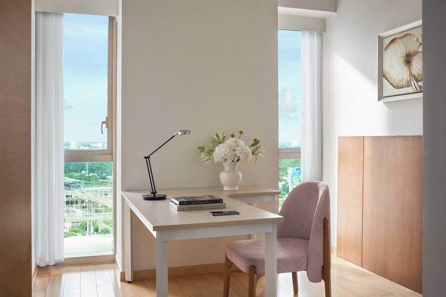 A modern office space with a white desk, a chair, a lamp, a vase with flowers, and a city view.