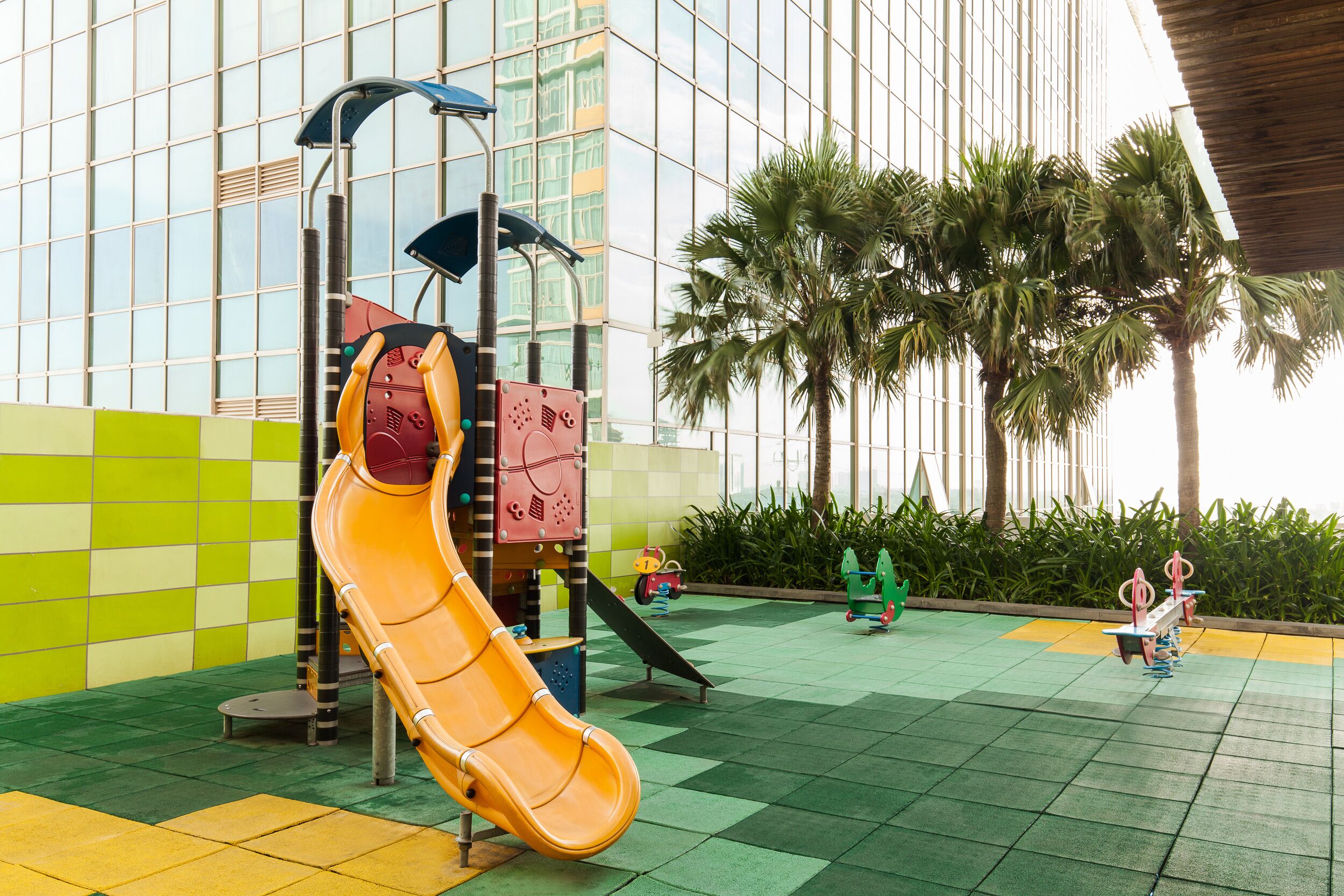 A playground with a yellow slide and climbing structures, set against a modern building with large windows and palm trees.