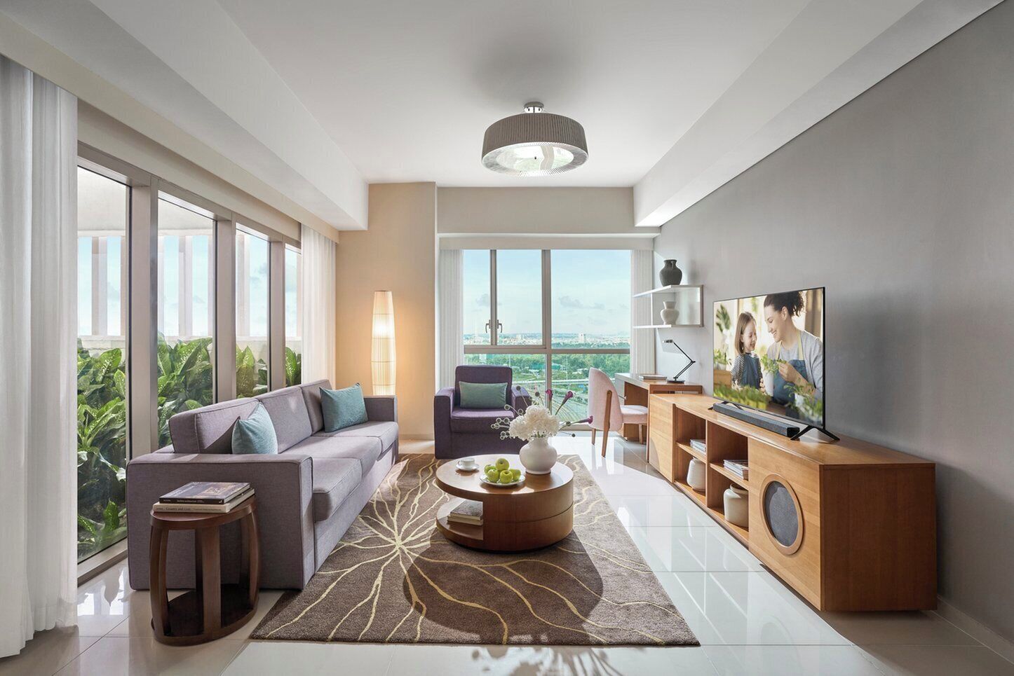 A modern living room with a sofa, coffee table, TV, and large windows offering a city view.