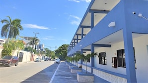 A street view with a modern building featuring a covered walkway, palm trees, and parked cars. - Fragata Departamentos (Puerto Morelos)