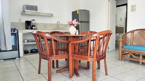 A compact kitchen with a microwave, stove, and refrigerator. There is a dining table with wooden chairs and a wicker chair with a blue cushion. - Fragata Departamentos (Puerto Morelos)
