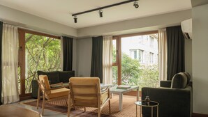 Habitación con sofá, sillón y una mesita con un libro. Amplias ventanas con cortinas, vistas al verdor exterior y una lámpara en el techo.