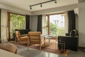 A room with a sofa, armchair, and a small table with a book. There are large windows with curtains, a view of greenery outside, and a ceiling light fixture. - Hub Porteño (Buenos Aires)