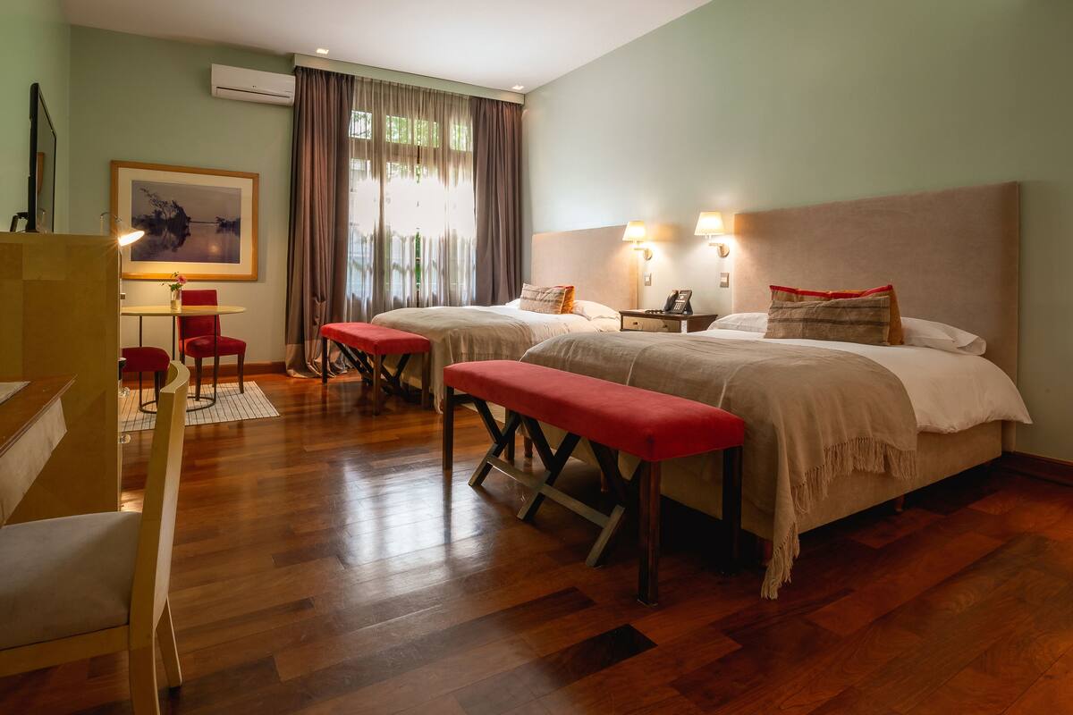 A hotel room with two beds, a desk with a chair, a bench, and a painting on the wall.