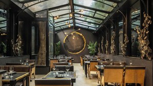 Un restaurante con techo alto, vigas a la vista y una lámpara circular central. Mesas dispuestas para comer, plantas decorativas y arte mural.