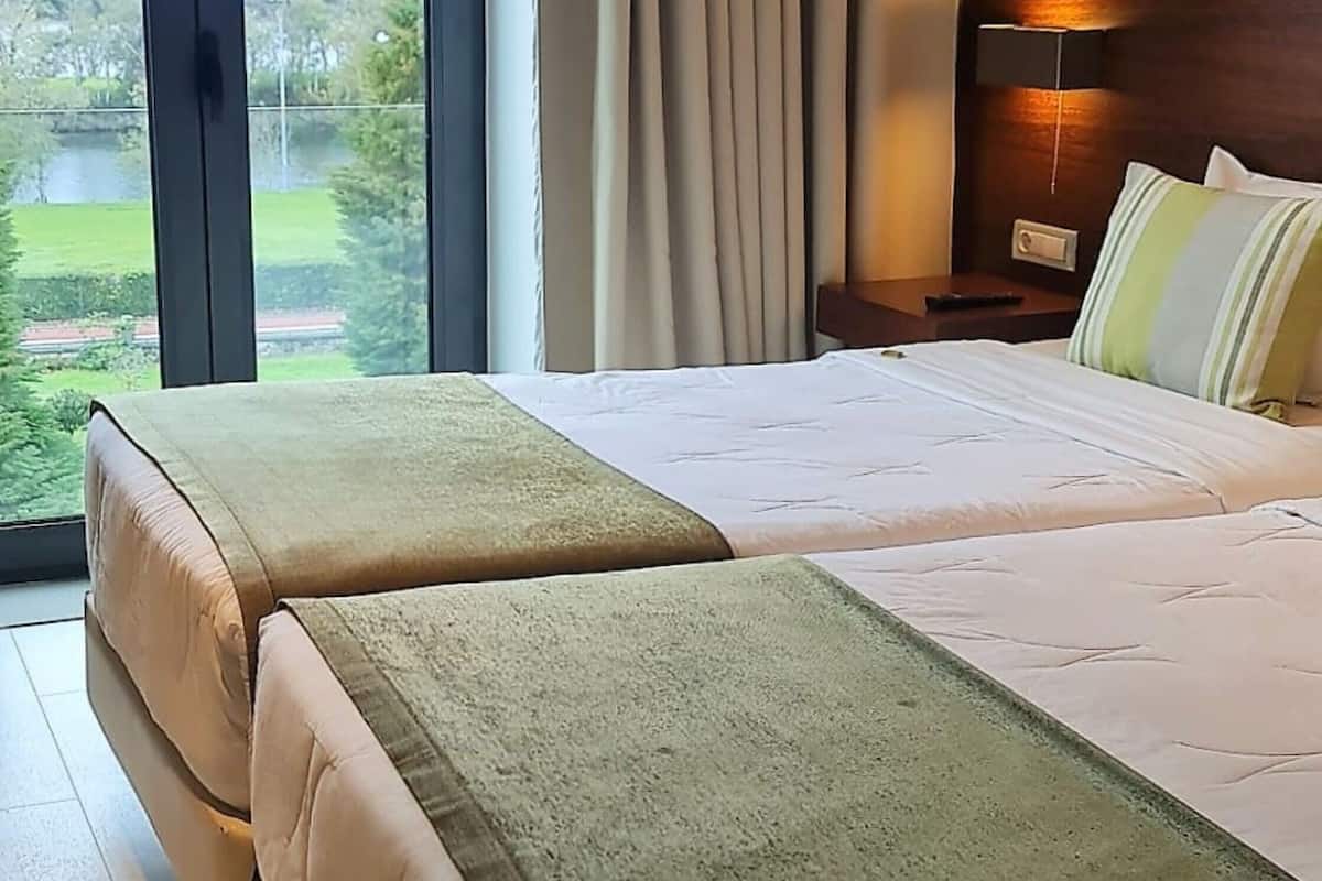 Une chambre d’hôtel avec deux lits, une grande fenêtre avec des rideaux, une table de chevet avec une lampe et une vue sur la verdure.