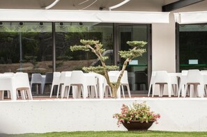 Uma área de estar moderna ao ar livre, com cadeiras de plástico branco e uma bonsai em primeiro plano.