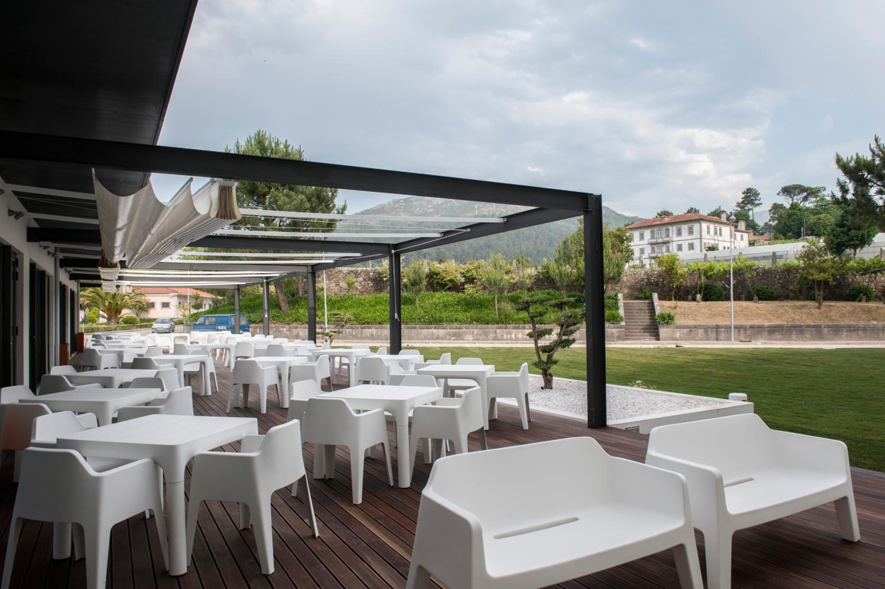 An outdoor patio with white plastic tables and chairs, a wooden deck, and a view of a building and greenery in the background.