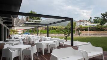 An outdoor patio with white plastic tables and chairs, a wooden deck, and a view of a building and greenery in the background.