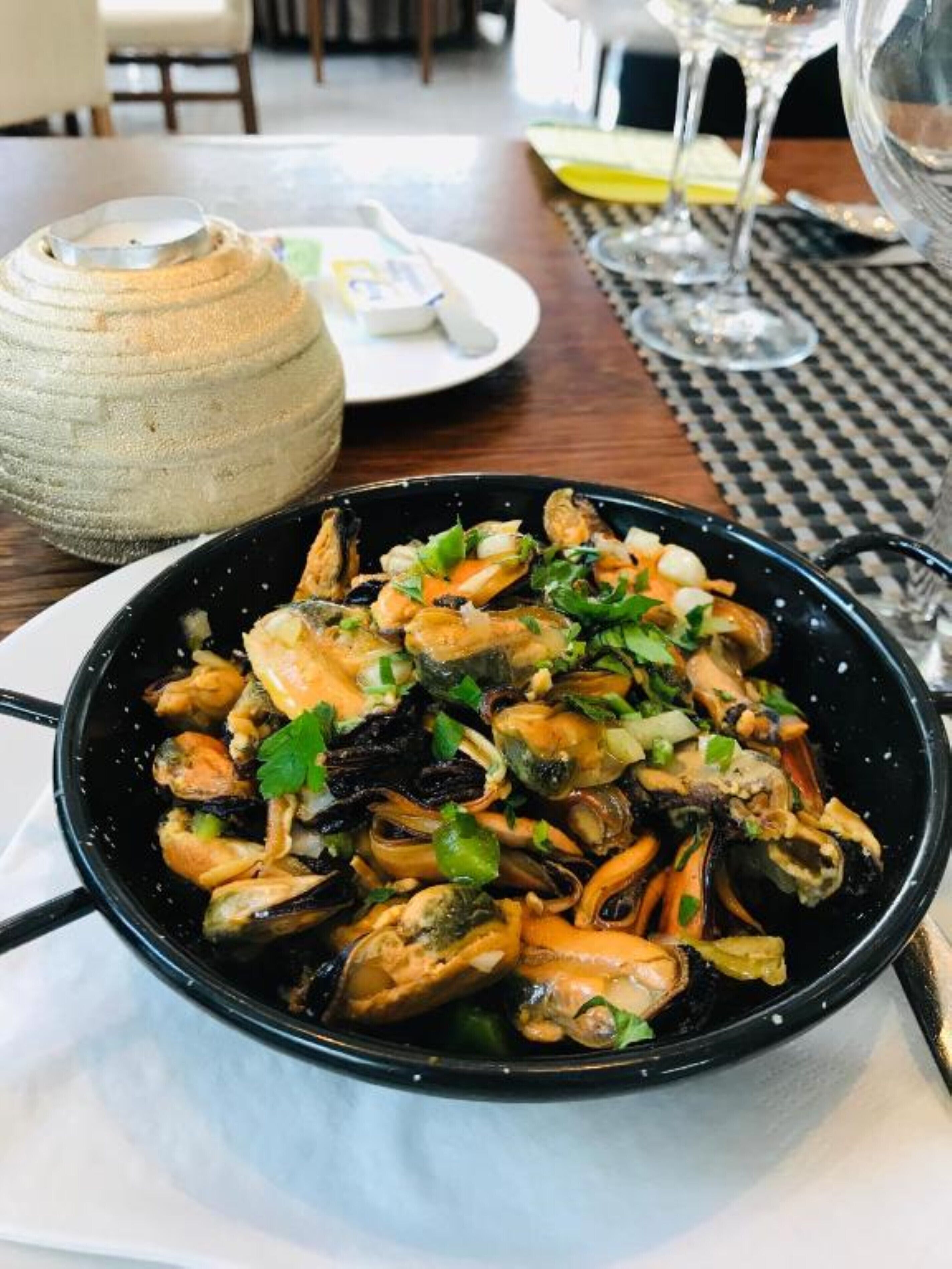 A bowl of seafood dish with mussels and herbs, placed on a table with a woven basket and a glass of water in the background.