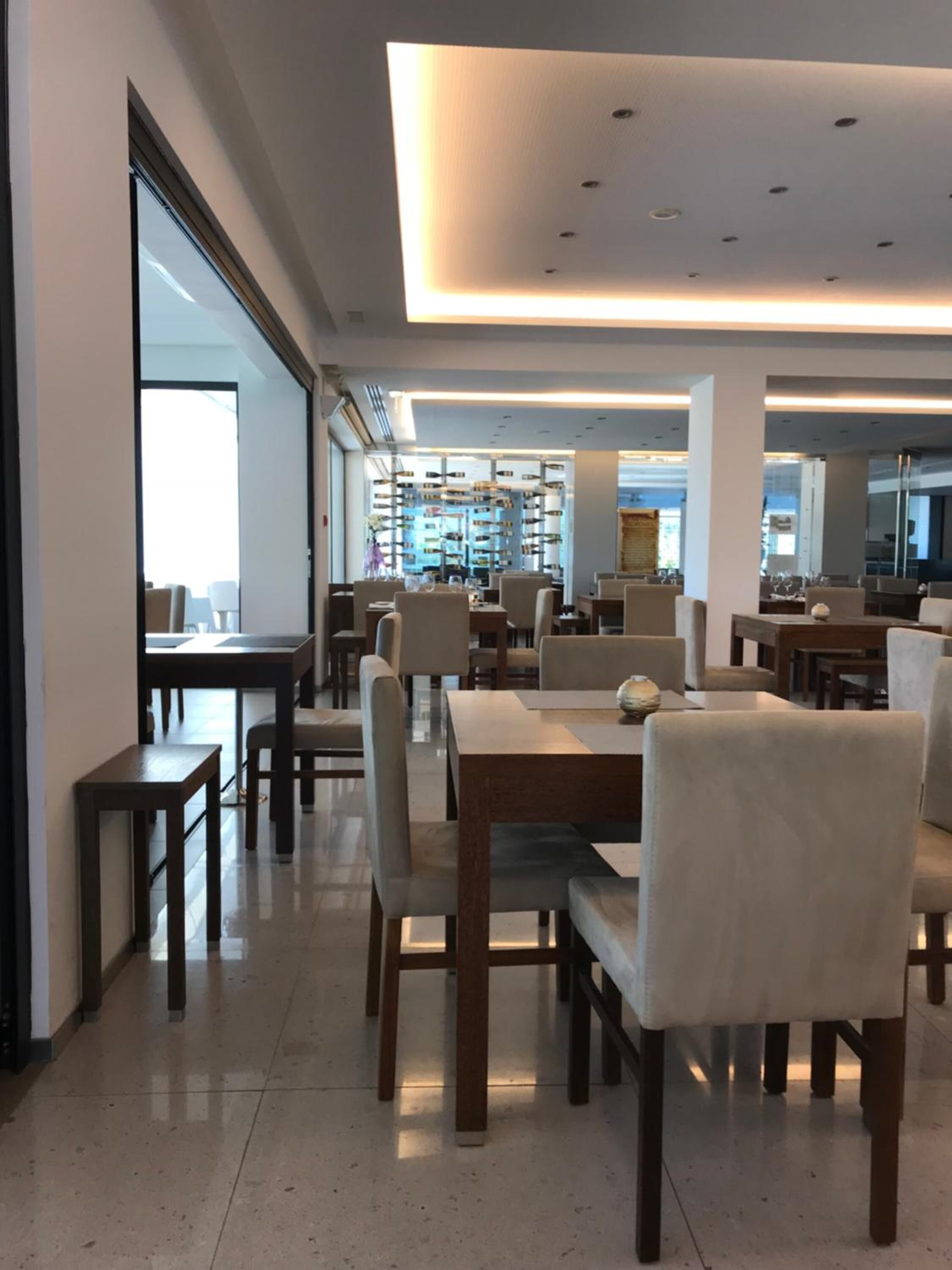 Un comedor espacioso con varias mesas y sillas, un diseño de techo moderno y vistas a una estantería para vinos al fondo.