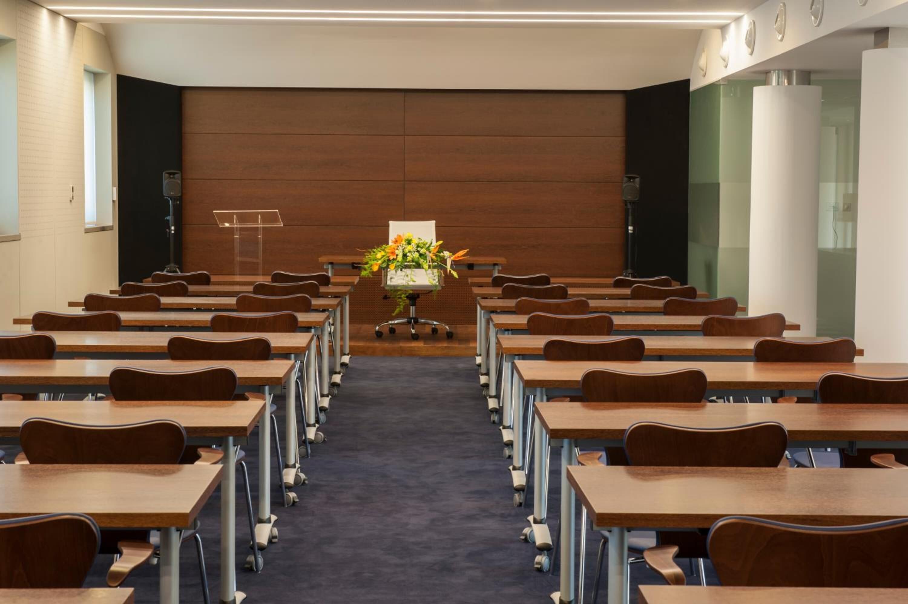 A conference room with rows of wooden tables and chairs, a podium at the front, and a floral arrangement on the podium.