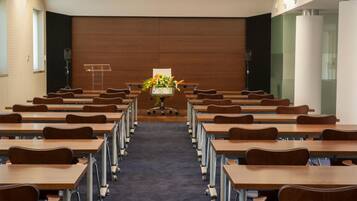 A conference room with rows of wooden tables and chairs, a podium at the front, and a floral arrangement on the podium.