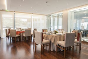 Uma sala de jantar com várias mesas preparadas para refeição, com piso de madeira e grandes janelas.