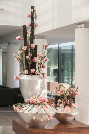 A modern interior with a large metallic vase containing pink flowers and branches, placed on a wooden platform.