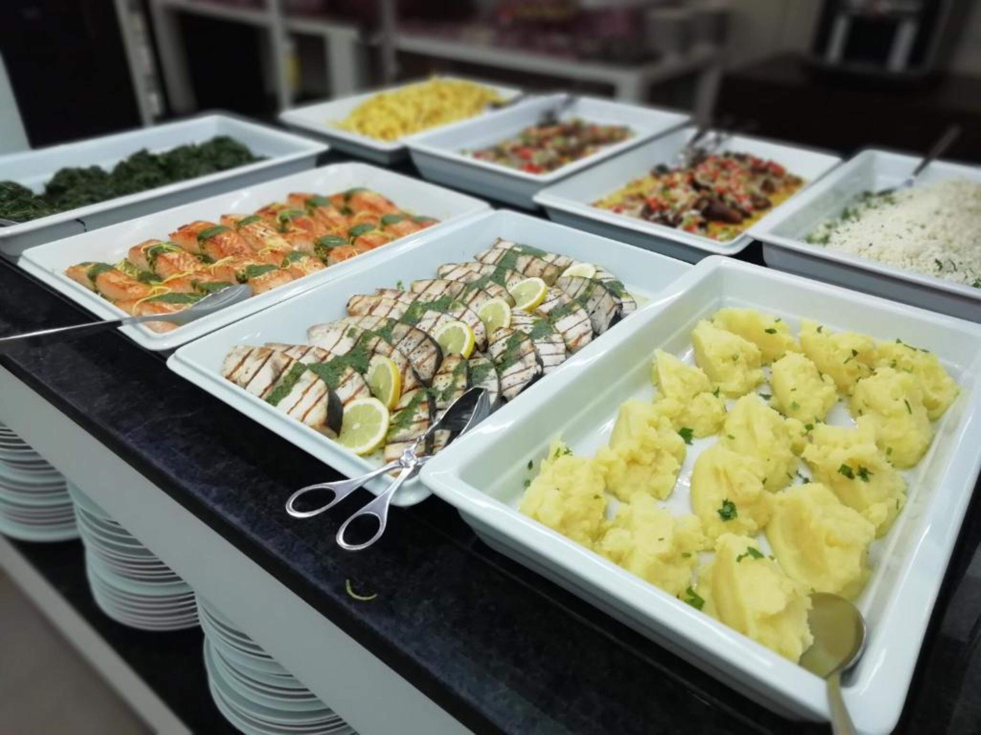Un bufé con diversos platos, incluyendo pescado a la parrilla, puré de papas y otros alimentos ya preparados.