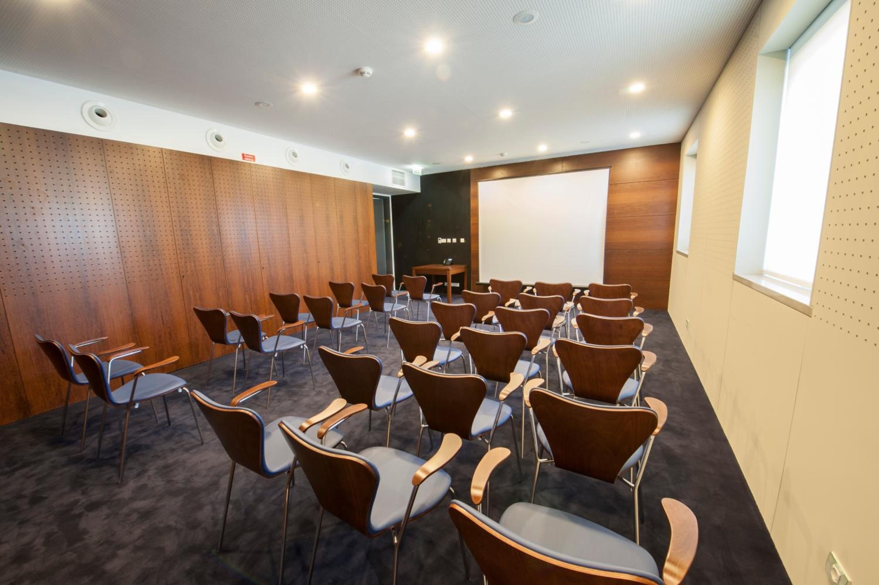 Un salón de conferencias con filas de sillas, una pantalla de proyector y una pared de madera decorativa.