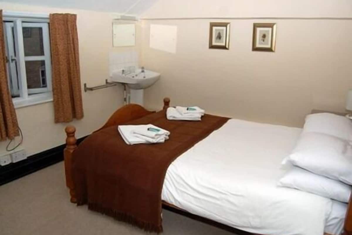 A bedroom with a bed, a wooden headboard, a brown throw blanket, white pillows, a white bedspread, a window with curtains, a wall-mounted sink, and two framed pictures.