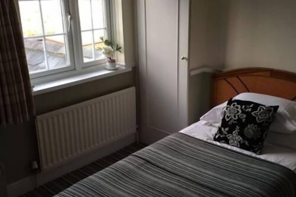 Une chambre à coucher avec un lit, une fenêtre avec des rideaux, un radiateur et une petite plante en pot posée sur le rebord de la fenêtre.