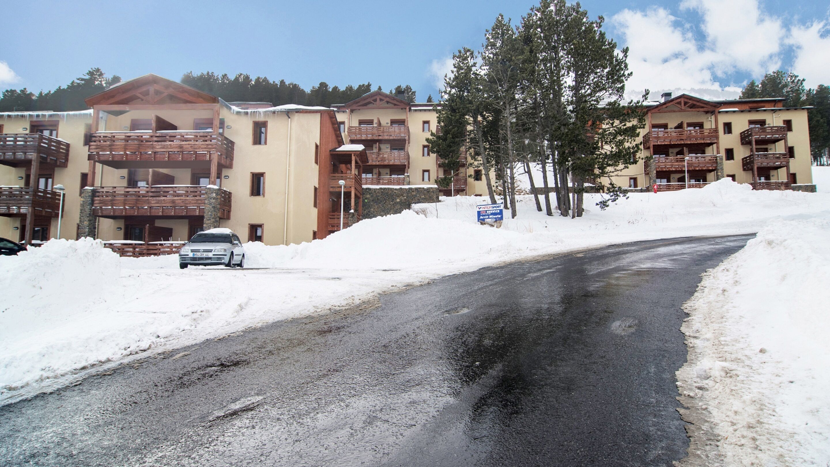 Une route enneigée avec une voiture garée sur le bord. On aperçoit des immeubles à plusieurs étages, dotés de balcons, ainsi qu'une enseigne.