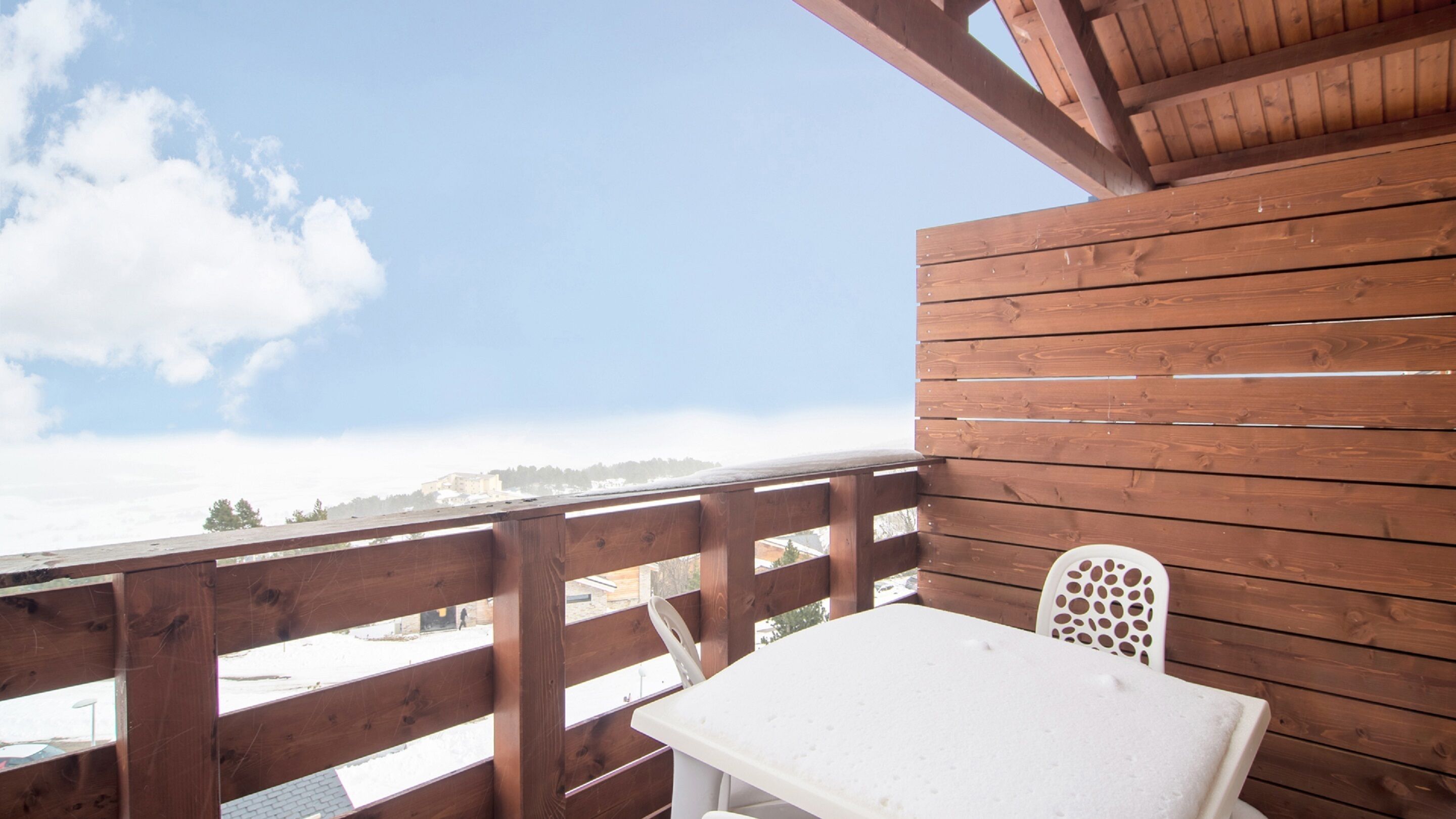 Une terrasse en bois avec une table et des chaises, d’où l’on peut admirer un paysage enneigé et des bâtiments.