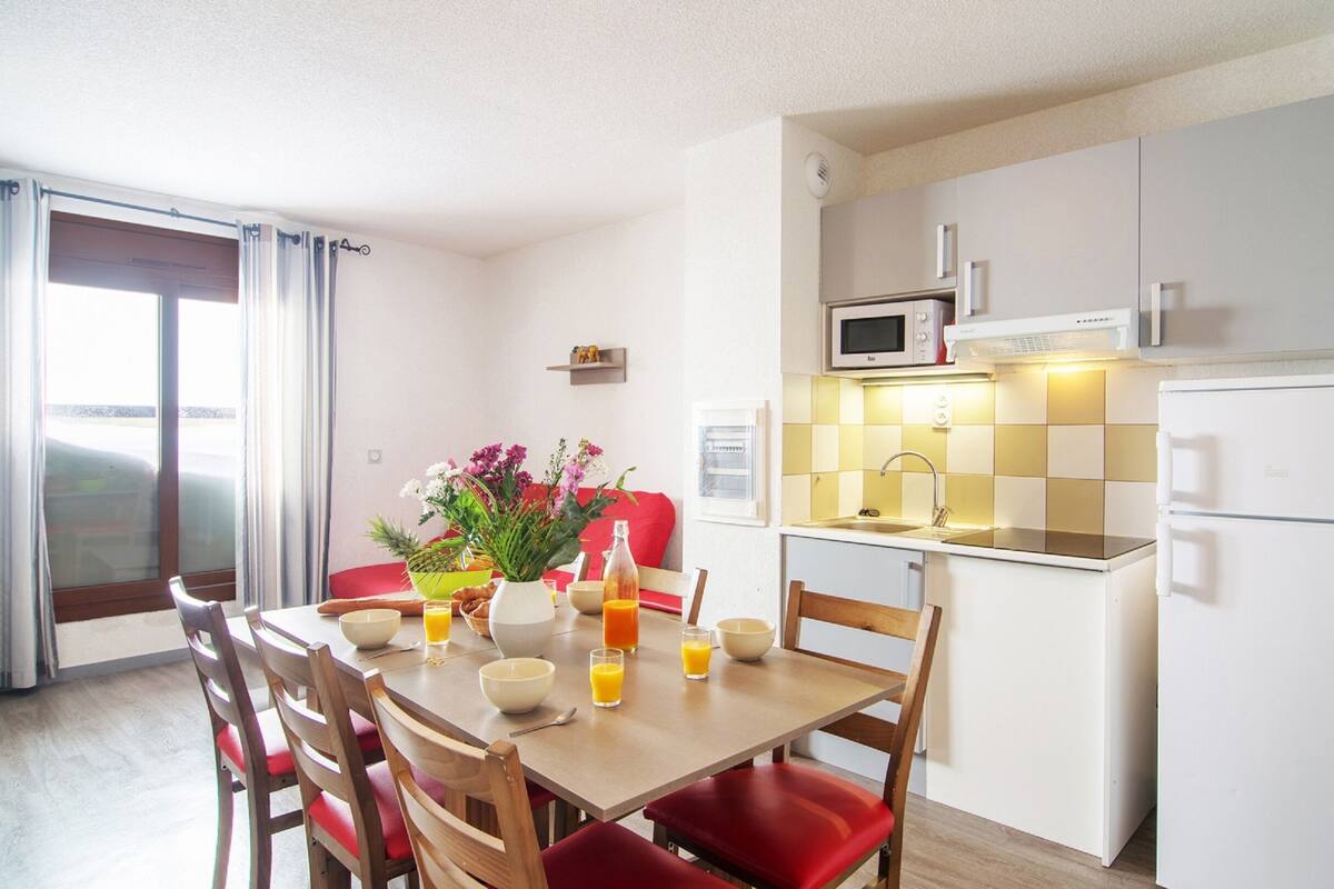 Une cuisine compacte avec des meubles blancs, un four à micro-ondes et un réfrigérateur. On y trouve une table à manger avec des chaises et un vase de fleurs.