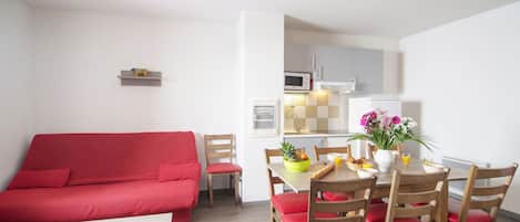 Un espace de vie compact comprenant un canapé rouge, une table à manger avec des chaises, un coin cuisine et une petite étagère ornée d’un objet décoratif.