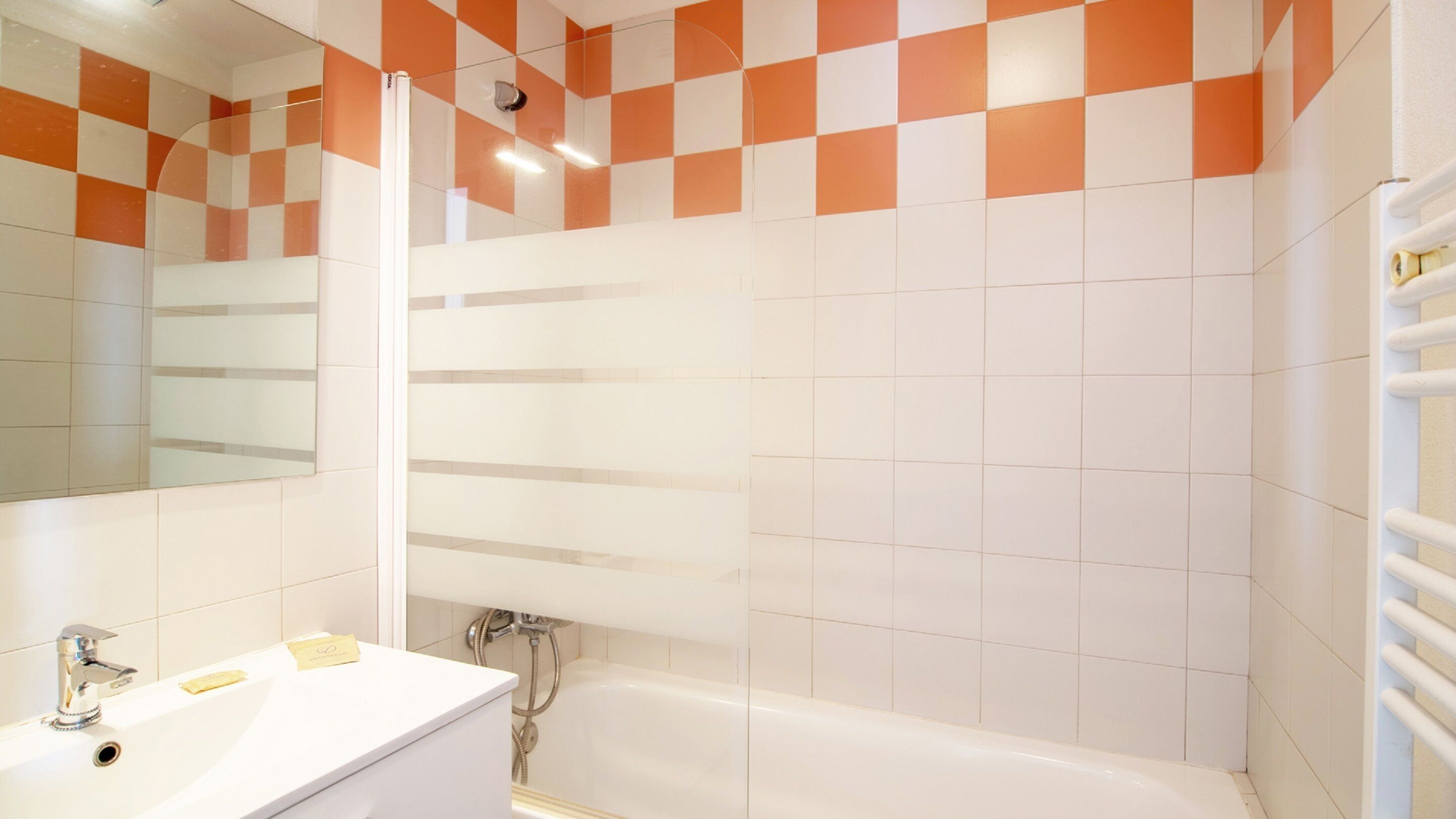 Een badkamer met een witte wastafel, een spiegel, een douche en oranje en witte geruitelde tegels.