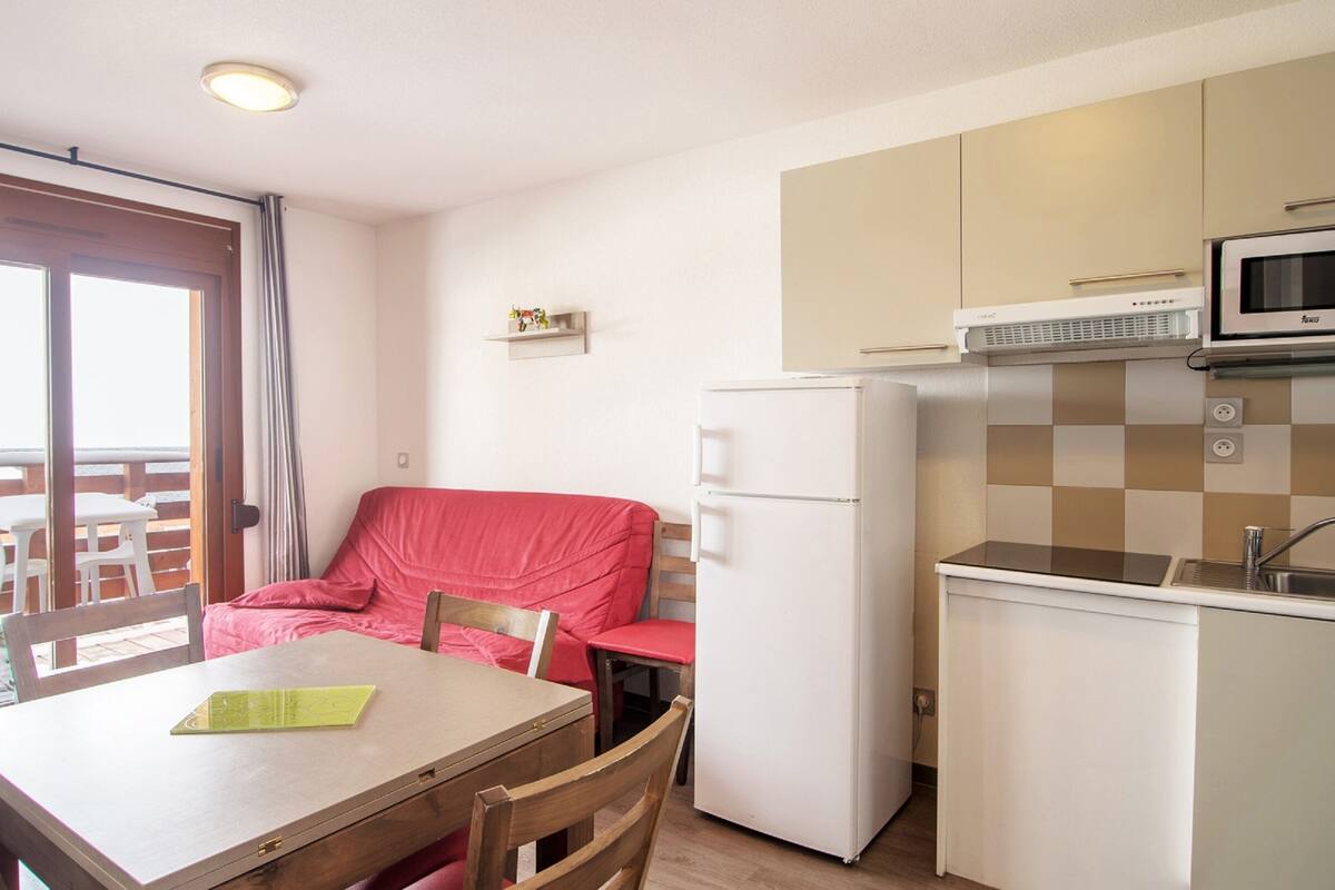 Une cuisine compacte équipée d’un réfrigérateur, d’un four à micro-ondes et d’un évier. On y trouve une table à manger avec des chaises et un canapé recouvert d’un tissu rouge.