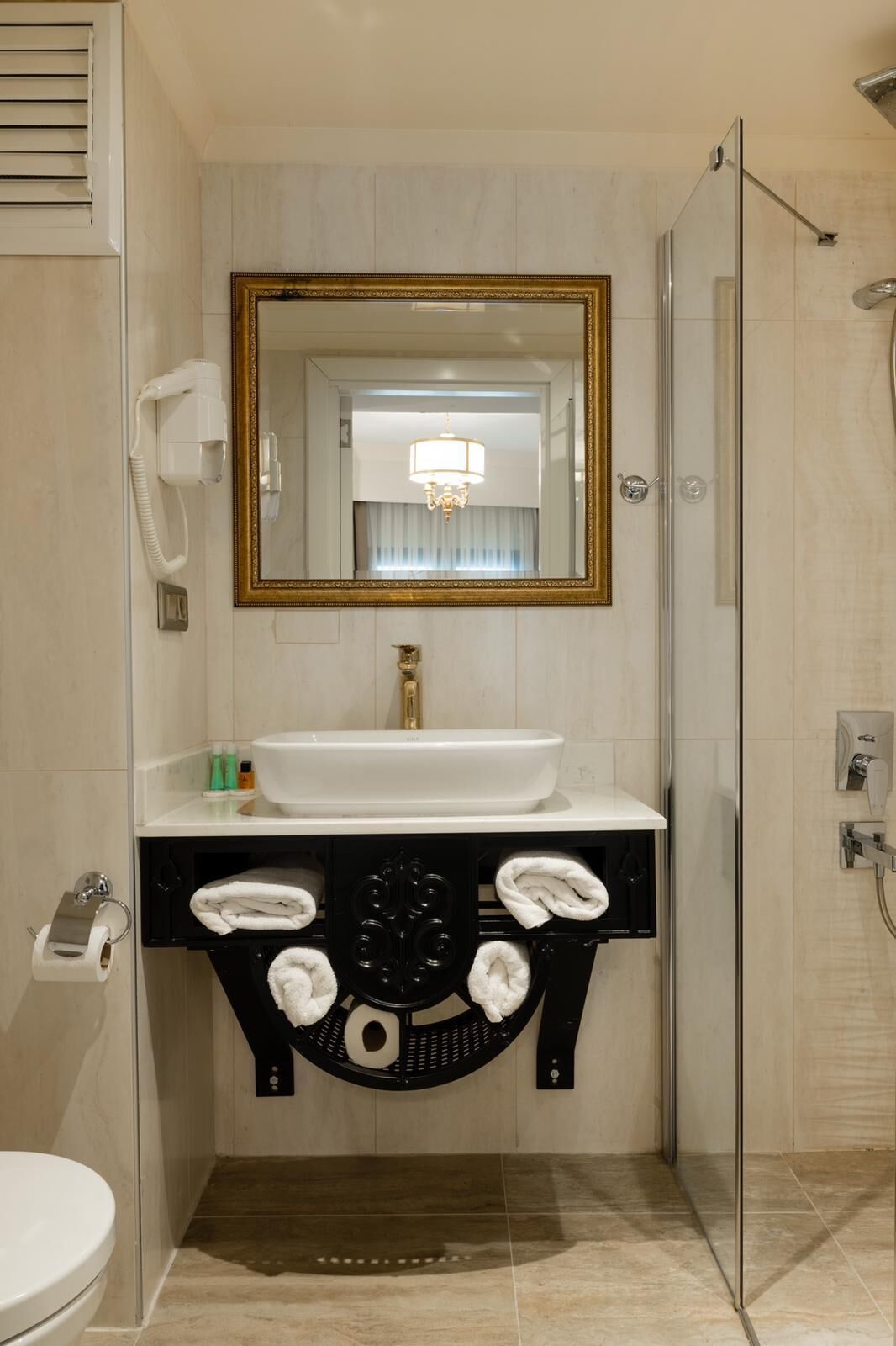 A bathroom with a white sink, a large mirror with a gold frame, a towel rack, and a shower area.