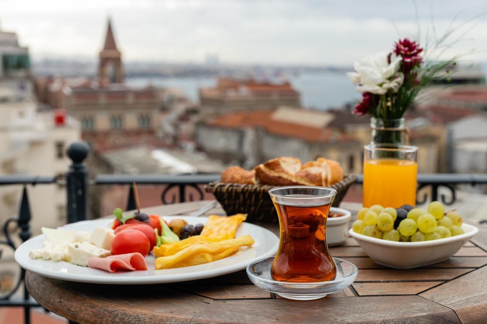 A table with a plate of food, a glass of tea, a bowl of grapes, and a glass of orange juice, with a city view in the background.