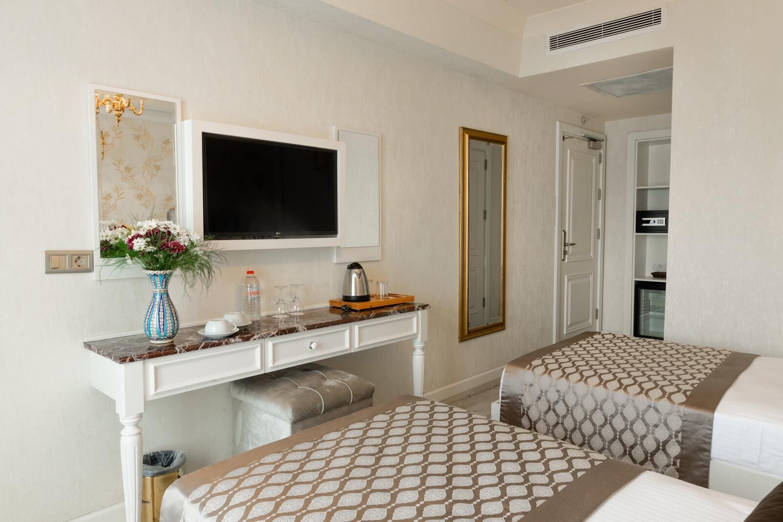 A hotel room with a bed, a desk with a TV, a mirror, and a minibar.