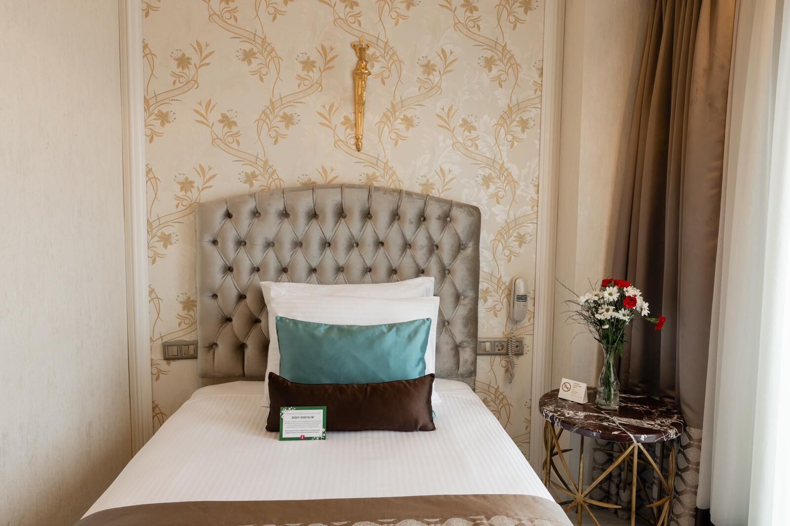 A neatly made bed with a tufted headboard, a bedside table with a vase of flowers, and a wall phone.