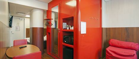 A compact hotel room with a red accent wall, a wooden floor, a small table with a chair, a microwave, and a red sofa.