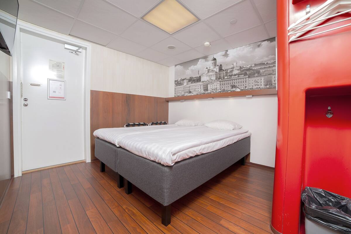 A bedroom with a bed, wooden flooring, a red cabinet, and a cityscape mural on the wall.