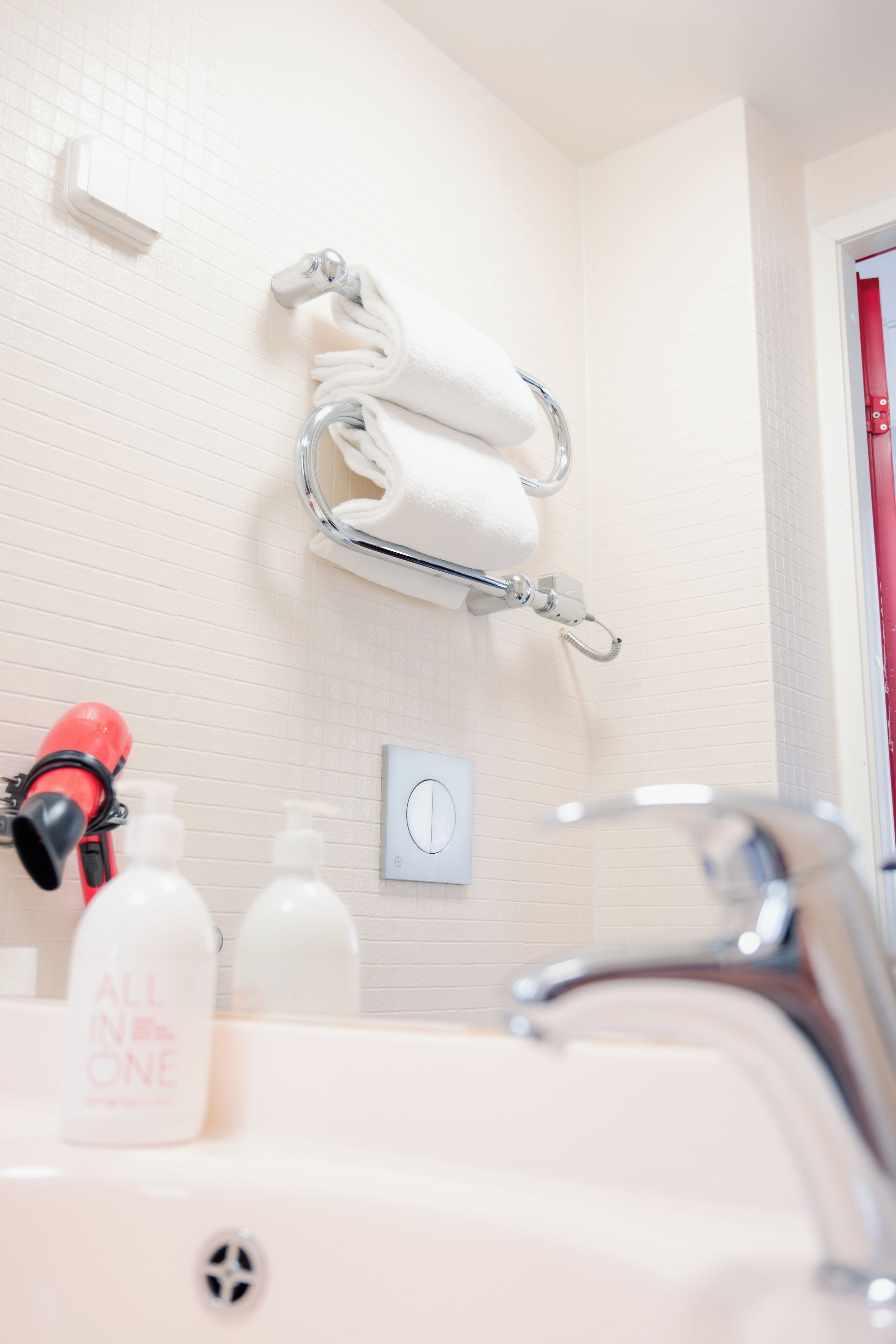 A bathroom with a white sink, a towel rack with two towels, a red hairdryer, and a bottle of All in One shampoo.