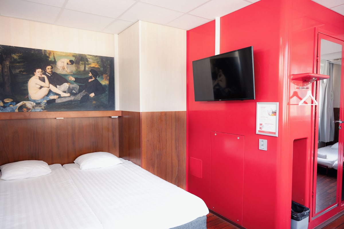 A hotel room with a bed, a painting on the wall, a television, and a red door.