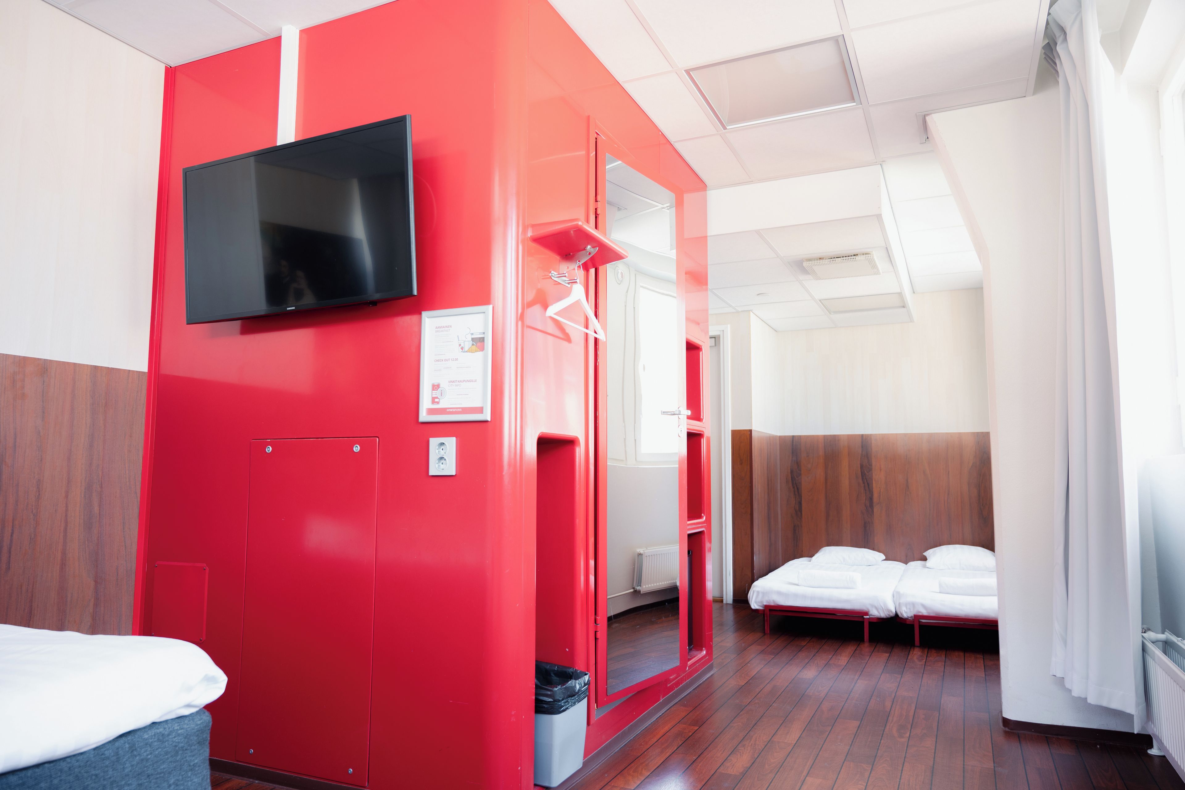 A hotel room with a red accent wall, a flat-screen TV, a mirror, and a wooden floor.