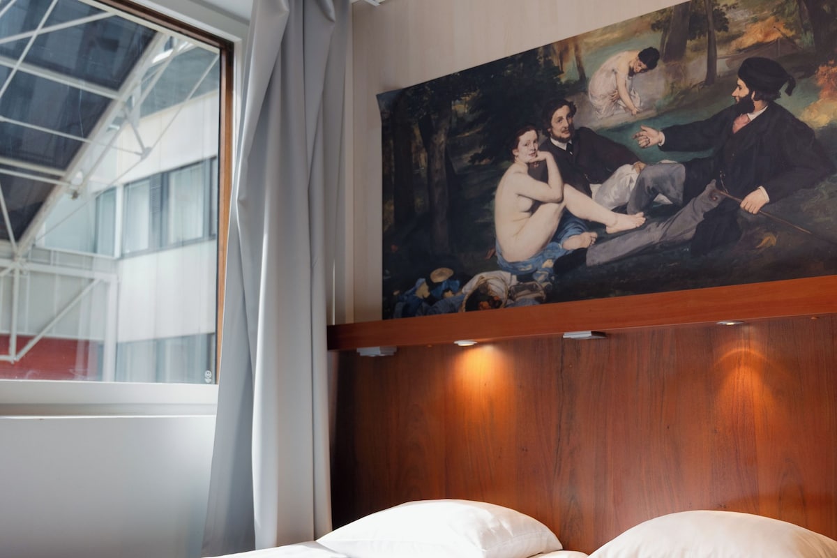 A hotel room with a bed, a painting on the wall, and a window with curtains.