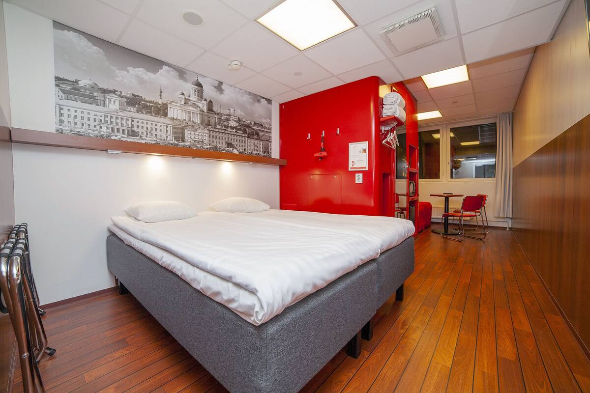 A hotel room with a bed, a red accent wall, a wooden floor, a window, and a view of a cityscape.