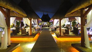 A covered outdoor dining area with tables and chairs, a wooden walkway, and thatched roofs. - Domaine des Alizées by Fine and Country (Grand-Baie)
