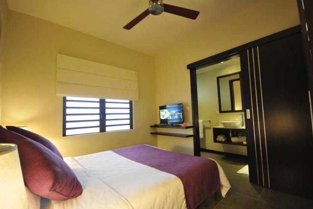 A hotel room with a bed, a television, a window with blinds, a ceiling fan, and a bathroom visible through an open door.