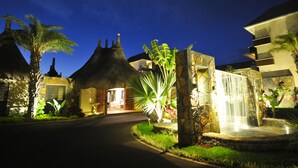 A resort entrance with a thatched-roof building, a stone wall with a waterfall, and lush greenery. - Domaine des Alizées by Fine and Country (Grand-Baie)