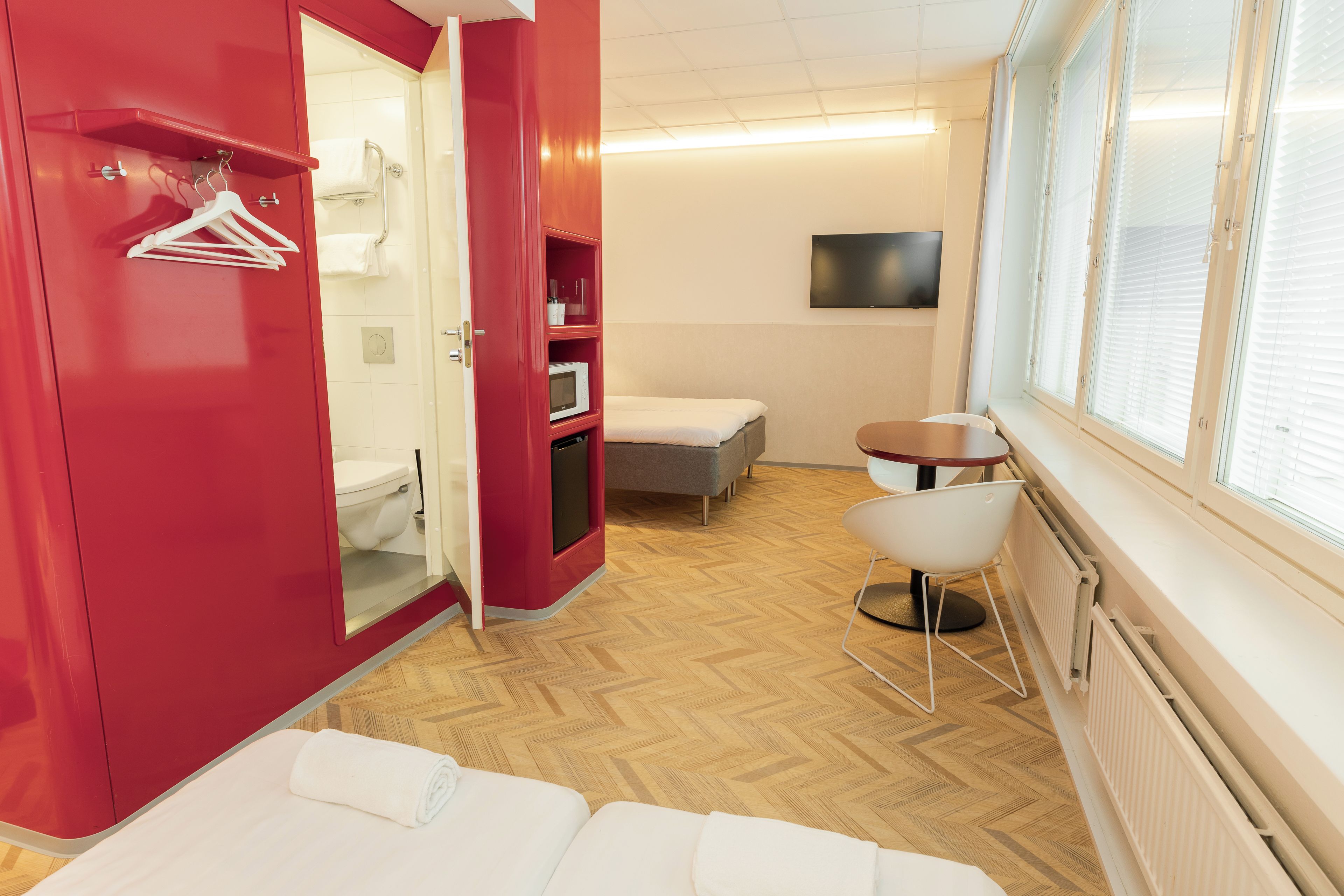 Ett hotellrum med en röd garderob, en dusch, en mikrovågsugn, en soffa, ett litet bord och en tv.