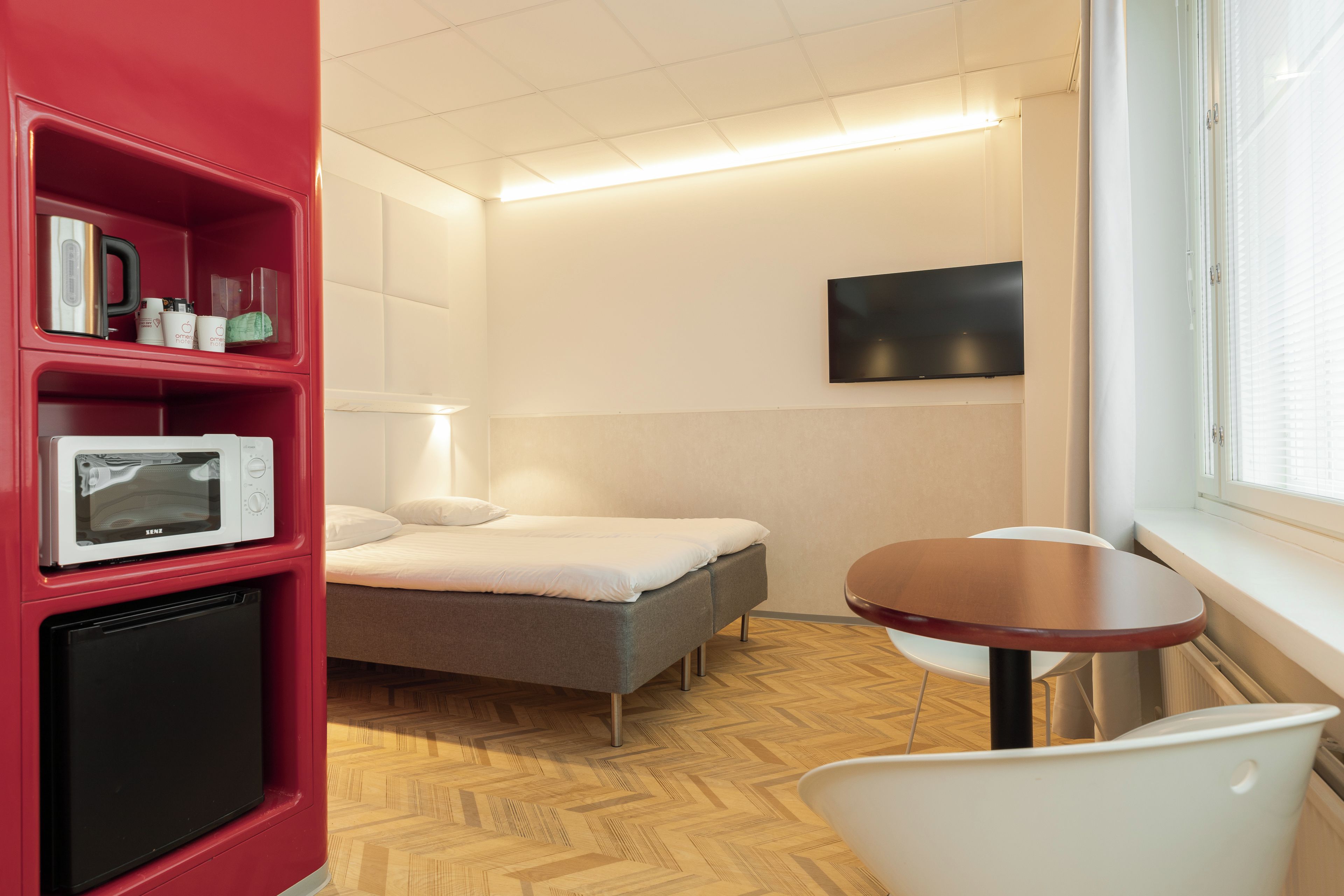 Habitación de hotel compacta con cama, una pequeña mesa, televisor montado en la pared y kitchenette con microondas.