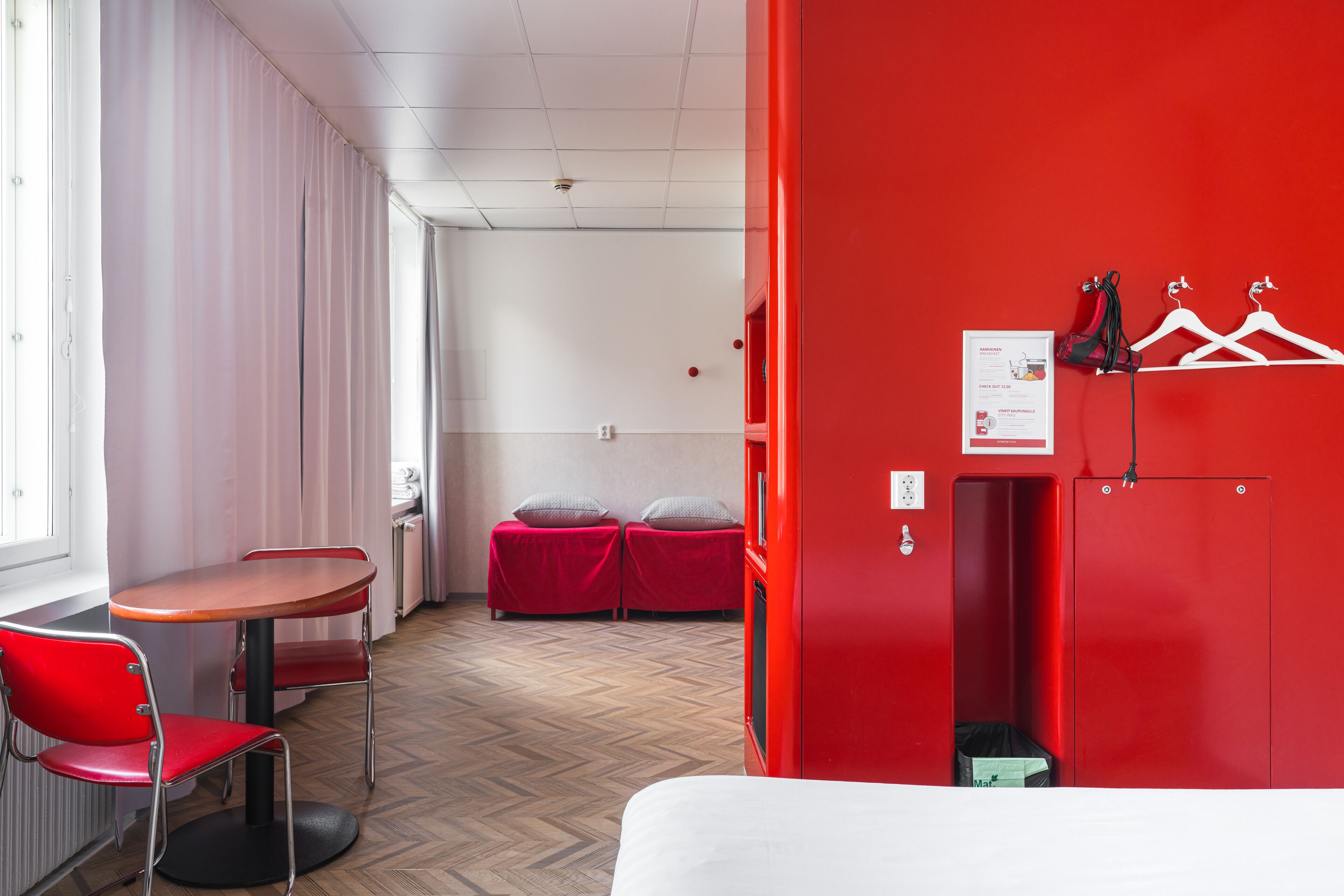 A hotel room with a red wardrobe, a bed, a table with a chair, and a small seating area with cushions.
