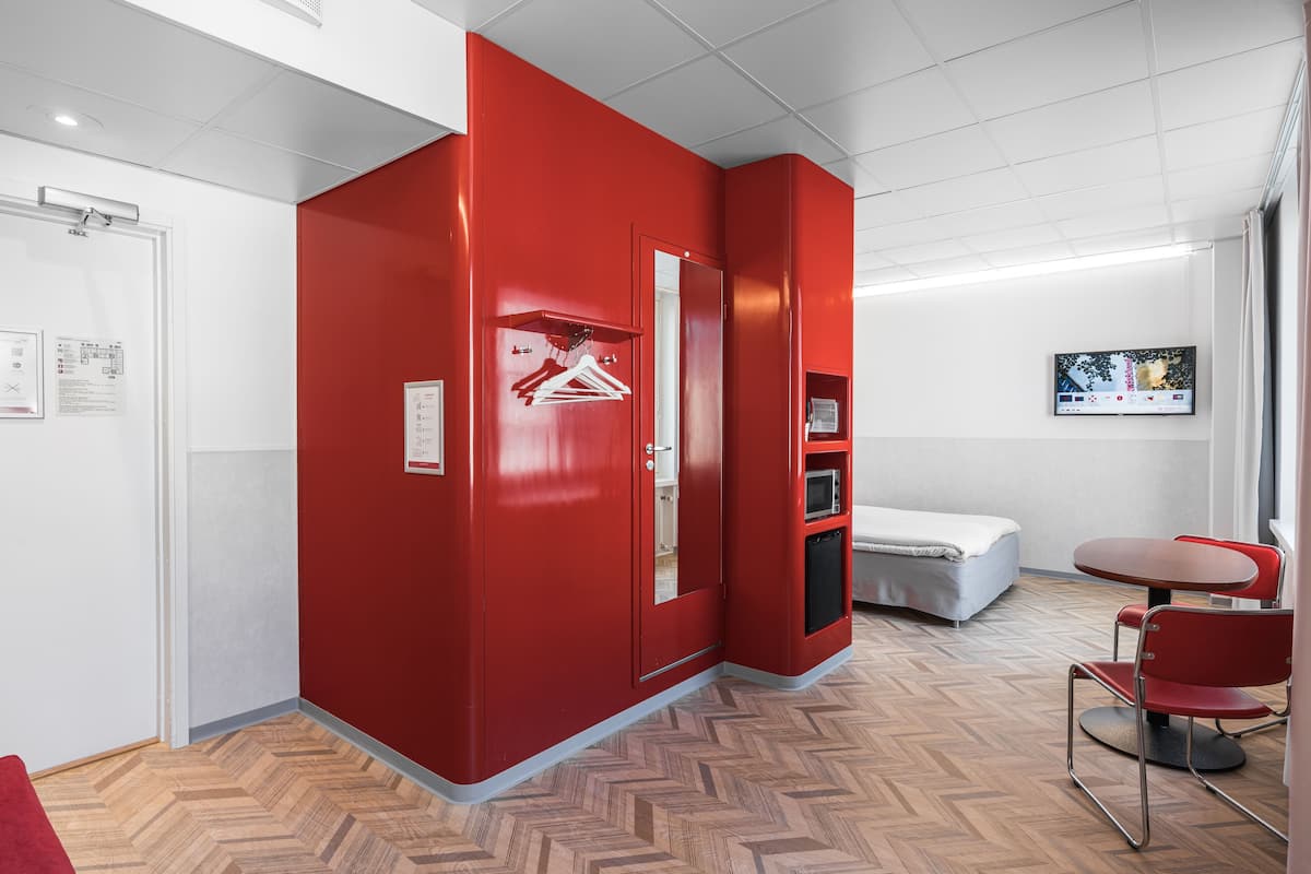 A hotel room with a red accent wall, a bed, a small table with chairs, and a coat rack.