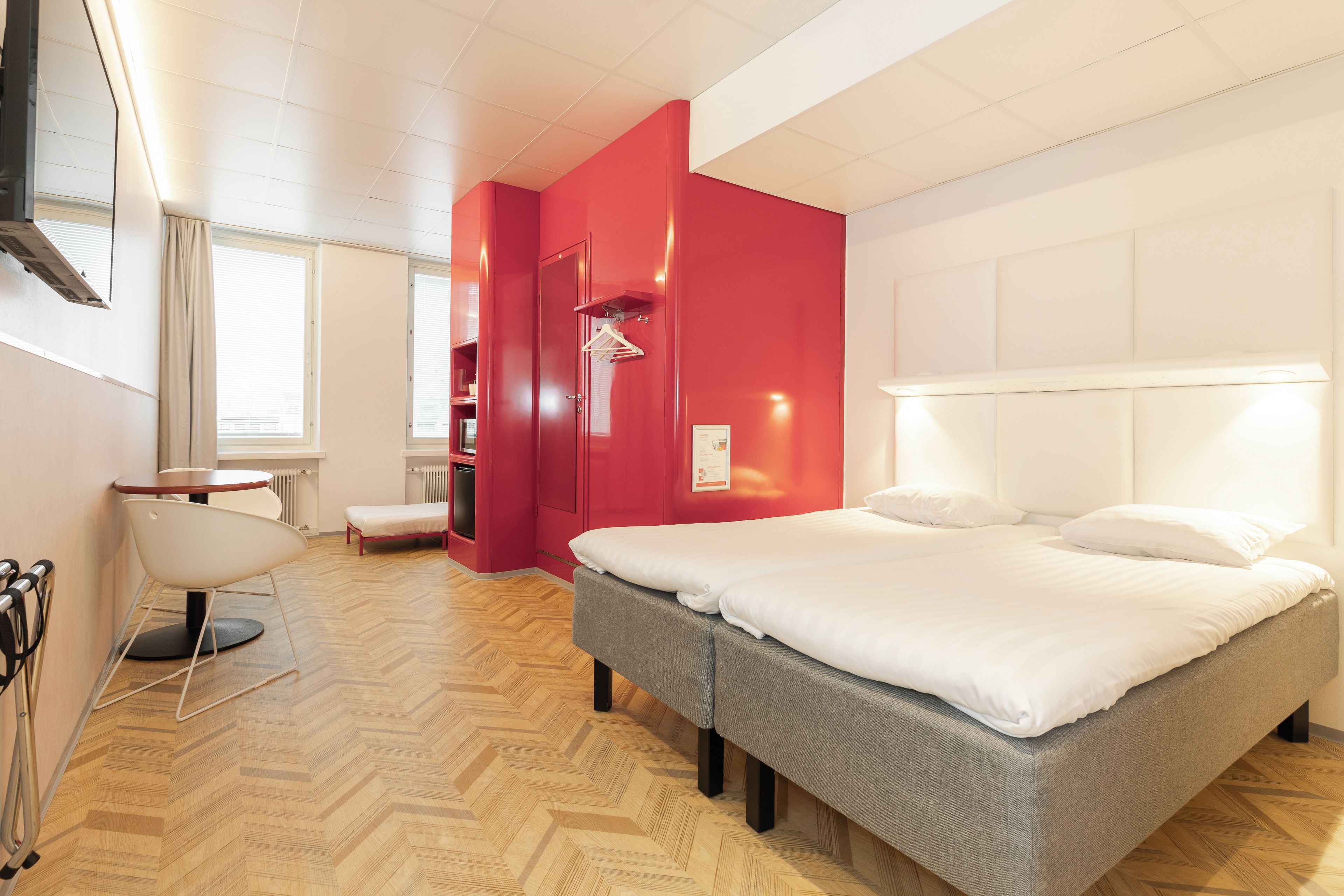 A hotel room with a bed, a chair, a small table, a radiator, and a window with blinds.
