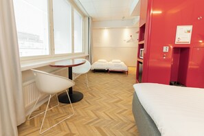 Une chambre d’hôtel avec un lit, une table ronde, deux chaises, une fenêtre avec des stores et un mur rouge sur lequel est affiché un avis.